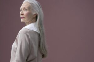 A sophisticated senior woman with long gray hair stands in profile against a solid background.