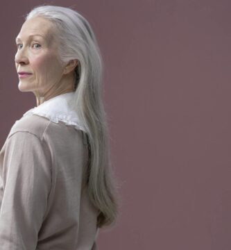 A sophisticated senior woman with long gray hair stands in profile against a solid background.