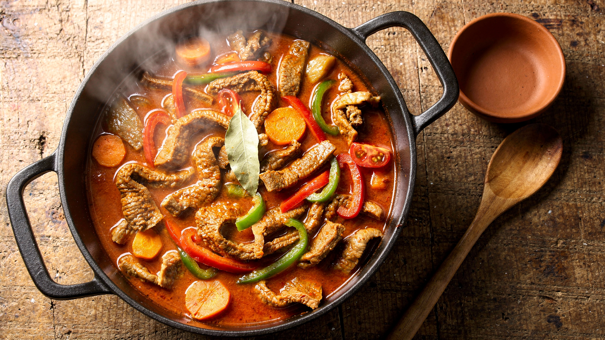 Olla de hierro con mondongo guisado en salsa roja espesa, con rodajas de zanahoria, tiras de pimentón rojo y verde, tomate y hojas de laurel